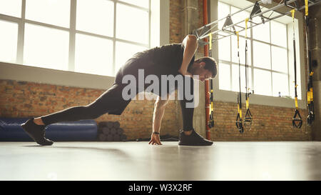 Preparing his muscles. Side view of young sporty man in sportswear stretching his legs while standing at gym. Workout concept. Professional sport. TRX Training. Stock Photo