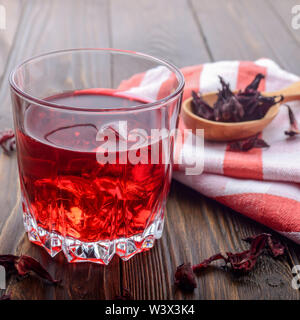 Closeup view at glass of tea with ice and spoon of dry hibiscus petals on wooden table background Stock Photo