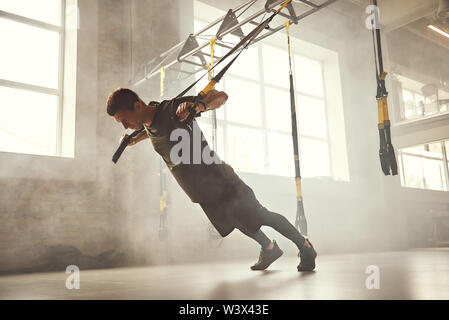 Perfect push-up. Side view of young and strong athletic man in sportswear doing push ups with trx fitness straps at gym. Fitness. TRX Training. Workout concept Stock Photo