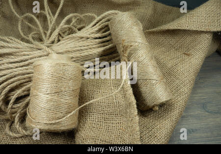 Two spools of burlap threads or jute twine and coil of linen rope on sackcloth fabric in close-up on grey background Stock Photo