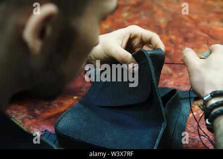 Craftsman intently works with leather. Tanner sews leather goods. Making handmade stuff. Male hands with needle and thread. Man working with needle. Stock Photo