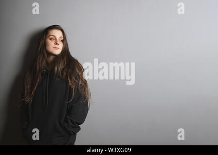 sad young goth woman wearing black sweater leaning against wall with her hands in pockets and looking to side in contemplation - gray background with copy space - adolescence and depression concept. Stock Photo