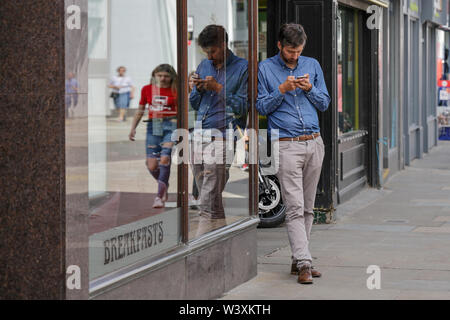 A handsome young man checks his smart phone in a seemingly deserted street, leaning against a shop window which reflects him and the busy street Stock Photo