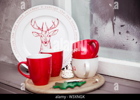 Red cups and a plate with painted deer stand on a wooden stand on the windowsill Stock Photo