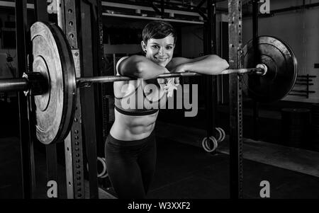 Black and white portrait of an attractive female athlete with strong abs. The athletic and smiling short-haired woman is leaning against the barbells. Stock Photo
