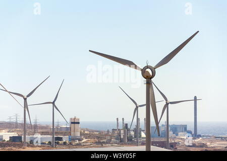 White turbines at wind farm with power station and sea in background, arid landscape on sunny summer day, sustainable renewable electricity concept Stock Photo