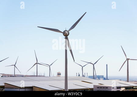 White turbines at wind farm with power station and sea in background, arid landscape on sunny summer day, sustainable renewable electricity concept Stock Photo