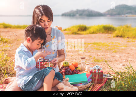 Beautiful Asian mother and son doing picnic and opening gift box from surprise in Birthday party on meadow near lake and mountain. Holiday and Vacatio Stock Photo