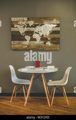 Vertical shot of two chairs and a table near a grey wall with a wooden world map on it Stock Photo