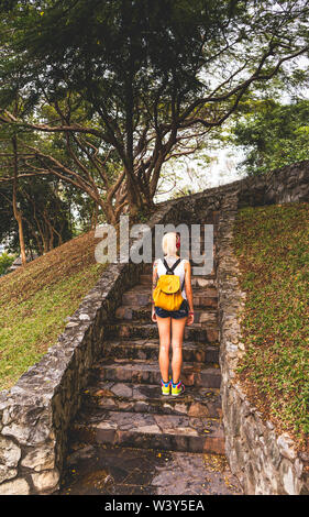 Blonde in denim shorts standing in the park on the stone steps. Wearing white jersey, orange backpack. Hands with tattoos. Asia travel Stock Photo