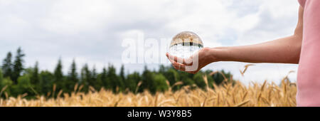 Woman holding crystal ball in her hand standing in nature with fields and sky reflecting in the ball in a conceptual image. Stock Photo