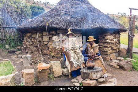 Basotho cultural village rural scene with two black African men dressed in traditional animal skins or hides outside a stone thatch rondawel or hut Stock Photo