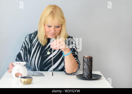 Middle age beautiful woman sits near a fortune teller desk, holding and using black pendulum on silver chain. Mystical psychic, clairvoyance and divin Stock Photo