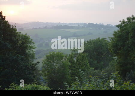 View from a hillside across trees towards open hilly countryside with summer sun low in cloudy sky The Doward South Herefordshire England UK