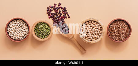 Flat lay in bowls a variety of legumes, red and white beans, chickpeas, mung beans on pastel pink background, banner. View from above