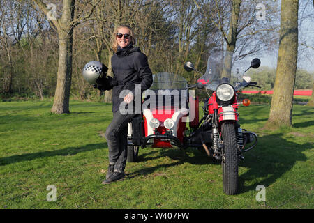 Young woman laughing and standing near a red motorcycle in the park. Stock Photo