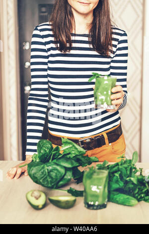 A brunette girl holds a glass with a ready smoothie in her hand, on the table in front of her is another glass with a ready smoothie, spinach foliage, Stock Photo