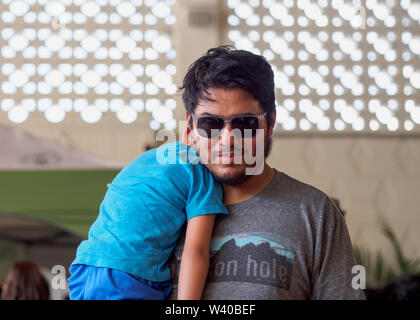 Dark haired man with short facial hair and sunglasses holds a small boy sleeping on his shoulder. 'Loca for Local' event Corpus Christi, Texas USA. Stock Photo