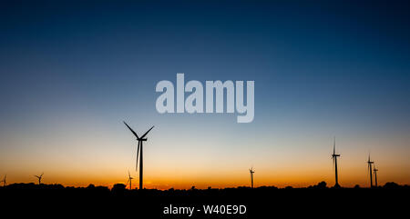 Black silhouettes of several wind turbines against orange and blue sky at dusk. Stock Photo