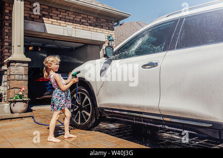 Cute preschool little Caucasian girl washing car on driveway in front house on sunny summer day. Kids home errands duty chores responsibility concept. Stock Photo