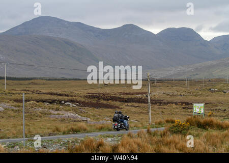 Two people on a motorbike ravelling on a road through bog land in County Galway towards Connemara with the mountains or Twelve Bens of Connemara ahead. Stock Photo