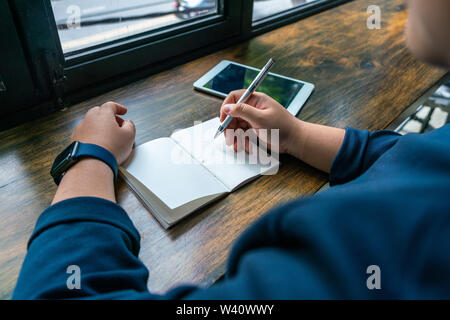 Woman holding pen and writing on notebook next to tablet Stock Photo