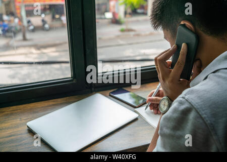 Asian man talking on cellphone and taking notes on table Stock Photo