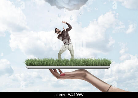 Female hand holding small man in casual clothes protecting himself from rainy cloud on green grass model with blue sky background. Digital art. People Stock Photo