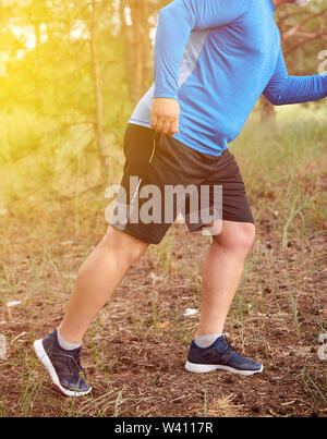 adult plump athlete in blue uniform, sneakers and black shorts runs through the middle of the woods on a summer evening,  concept of outdoor sports an Stock Photo