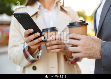 Cropped photo of office workers man and woman in formal wear holding takeaway coffee while using mobile phone on city street Stock Photo