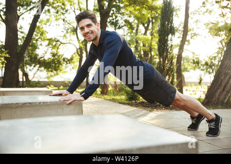 Portrait of happy athletic man dressed in sportswear doing push-ups and stretching his body while working out in sunny green park Stock Photo