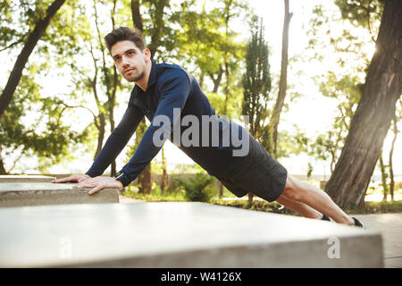 Portrait of focused athletic man dressed in sportswear doing push-ups and stretching his body while working out in sunny green park Stock Photo