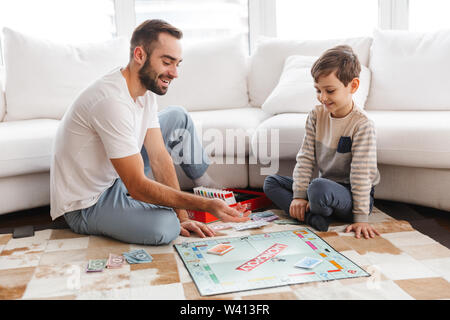 Cheerful young father playing monopoly with his little son while sitting on a floor at the living room Stock Photo