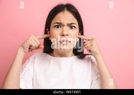 Portrait closeup of sad teenager girl in casual wear looking at camera and plugging her ears with fingers isolated over pink background Stock Photo