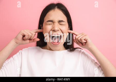 Portrait closeup of scared teenager girl in casual wear screaming and plugging her ears with fingers isolated over pink background Stock Photo