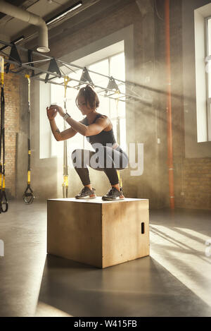 Challenging myself. Side view of athletic woman in sportswear doing squat while standing on wooden box at gym. Professional sport. TRX Training. Workout Stock Photo