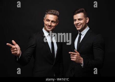 Portrait of smiling handsome two businessmen dressed in formal suit pointing fingers and looking aside isolated over black wall Stock Photo
