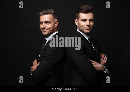Portrait of smiling handsome two businessmen dressed in formal suit posing at camera back to back isolated over black wall Stock Photo