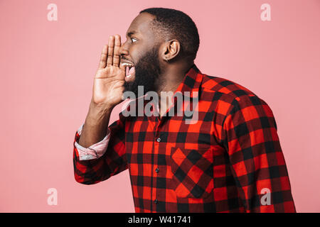 Portrait of an excited african man wearing plaid shirt standing isolated over pink background, shouting, looking away Stock Photo