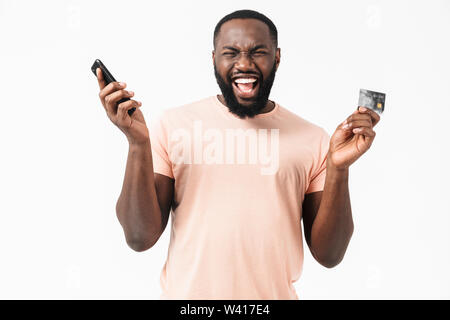 Portrait of a happy african man wearing t-shirt standing isolated over white background, showing credit card while using mobile phone Stock Photo