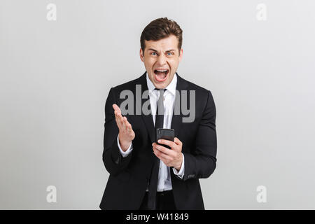 Image of angry yong businessman in formal suit typing on cellphone and screaming at camera isolated over gray background Stock Photo