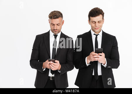 Image of successful serious two businessmen in office suits holding and typing on cellphones isolated over gray background Stock Photo