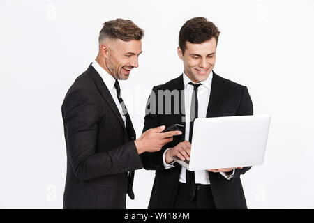 Image of two happy caucasian businessmen in office suits holding laptop and cellphones isolated over gray background Stock Photo