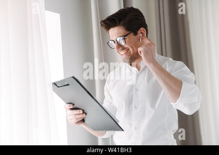 Handsome confident businessman standing at the window indoors, holding tablet, wearing wireless earphones Stock Photo