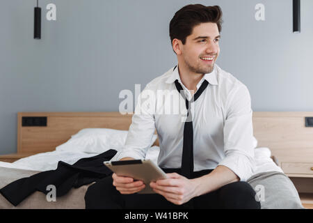 Photo of handsome smiling businessman wearing formal clothes using tablet computer while sitting on bed in hotel apartment Stock Photo