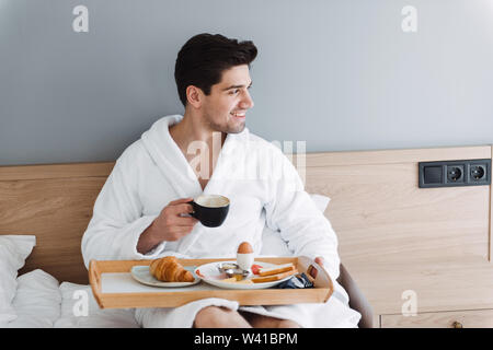 Photo of unshaven european man wearing white bathrobe having breakfast while sitting on bed in hotel apartment Stock Photo
