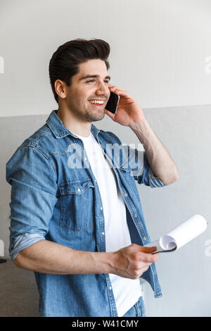 Attractive young man standing at the kitchen at home, holding clipboard, working, talking on mobile phone Stock Photo