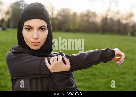 Portrait of sporty muslim woman dressed in religious black hijab stretching her arms while doing workout in green park outdoors Stock Photo
