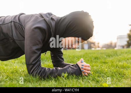 Image of a young concentrated muslim sports fitness woman dressed in hijab and dark clothes outdoors in green nature park make exercises plank. Stock Photo