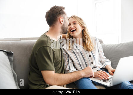 Image of a happy excited young loving couple sitting on sofa at home using laptop computer. Stock Photo